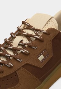 Baskets marron et beige avec des panneaux en daim texturé, des lacets marron et blancs réfléchissants, une languette beige perforée, et le logo Tommy Jeans sur le côté.