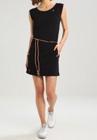 Frau in einem ärmellosen schwarzen Kleid mit weißen Punkten, einem dünnen braunen Gürtel und weißen Turnschuhen, die vor einem einfarbigen Hintergrund steht.