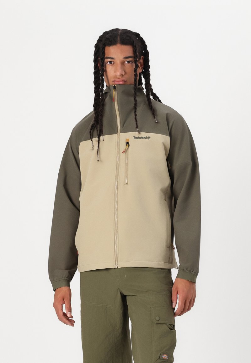 Beige og olivengrønn zip-jakke med høy krage, to sidelommer og Timberland-logo på brystet. Jevn stofftekstur.