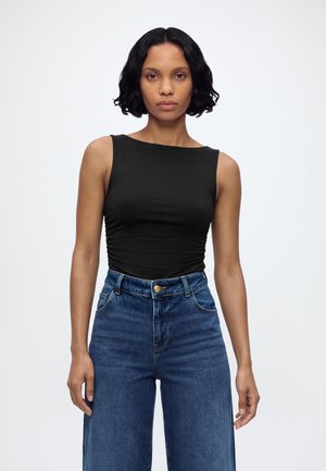 Mujer con cabello corto y rizado de color negro, que lleva una camiseta negra sin mangas y jeans azules de cintura alta, de pie sobre un fondo blanco.