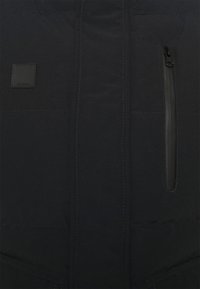 Chaqueta acolchada negra con un bolsillo con cremallera y una etiqueta cuadrada con logo. Presenta costuras en contraste y una textura suave.