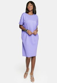 Liliowa sukienka z krótkimi rękawami, luźny krój i boczne kieszenie. Gładka faktura materiału, długość do kolan, zestawiona z przezroczystymi sandałami na kostkę.