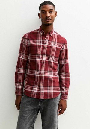 Un hombre está de pie contra un fondo simple, vistiendo una camisa de cuadros rojos y jeans grises, mirando directamente a la cámara con una expresión de confianza.