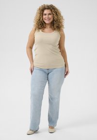Débardeur beige avec des ornements, associé à un jean droit bleu clair. Le mannequin se tient sur un fond neutre, portant des chaussures beiges.