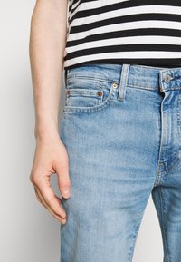 Vaaleansiniset farkut klassisella leikkauksella. Ominaisuuksiin kuuluvat viisi taskua, metallinen napitus ja hillitty haalistuminen. Raidallinen paita näkyvissä.