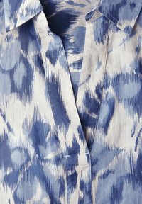 Camisa azul y blanca con un patrón abstracto texturizado. Presenta un cuello abotonado y una tapeta con un escote recto.