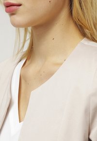 Gros plan sur le bas du visage et le cou d'une femme à la peau claire, aux cheveux blonds, portant un haut blanc à col en V et un blazer beige clair.