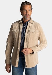Beige corduroy overhemd met een knoopsluiting aan de voorkant, twee borstzakken en lange mouwen. Gedragen over een donkerblauw overhemd met knopen.
