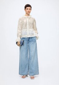 Femme portant un chemisier blanc transparent brodé, un jean bleu clair à jambes larges, tenant une pochette bleue et des sandales blanches à talons.