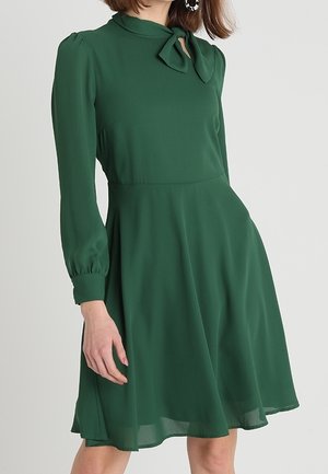 Vrouw die een groene jurk met lange mouwen, een hoge halslijn en een strik bij de kraag draagt, staand tegen een effen achtergrond.