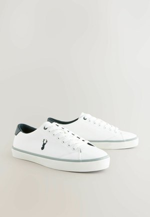 Valkoiset kanvaslenkkarit mustilla nahkakiinnikkeillä, joissa on logo sivussa, nauhakiinnitys ja kumipohja harmailla yksityiskohdilla.