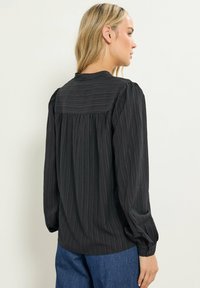 Blond kvinde iført en langærmet sort tekstureret bluse med samlet halsudskæring og mørkeblå jeans, der står sidelæns mod en ensfarvet hvid baggrund.