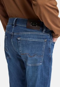 Ciemnoniebieskie dżinsy w kolorze denim o średnim stanie, z widocznym przeszyciem kieszeni oraz brązową skórzaną naszywką z tyłu z metalowym logo.