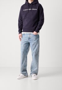Marinblå hoodie med vit "Calvin Klein Jeans"-logotyp, ihop med ljusblå löst sittande jeans och vita sneakers på en neutral bakgrund.