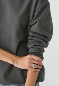 Sudadera gris con mangas enrolladas, con una textura suave. Un anillo de plata con gemas se lleva en la mano, reposando sobre el brazo.