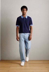 Marinblå stickad polo med ljusblå krage och manschetter, ihop med ljusblå lösa jeans och vita sneakers på ett trägolv.