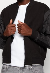 Zwarte bomberjack met gepocheerde zwarte nylon mouwen. Voorzien van een rits aan de voorkant, geribde manchetten en een gladde textuur. Gedragen over een wit shirt.