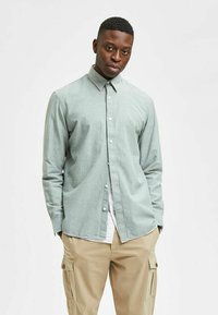 Lichtgroene blouse met kraag en lange mouwen, gemaakt van een gestructureerde stof, gedragen over een wit t-shirt en gecombineerd met beige cargo-broeken.