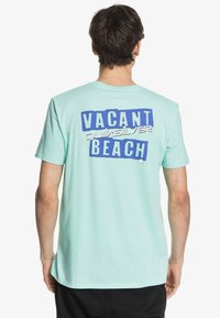 Muž otočený zády, oblečený v světle modrém tričku s nápisem "VACANT BEACH Quiksilver" v tučných modrých a bílých písmech na zádech.