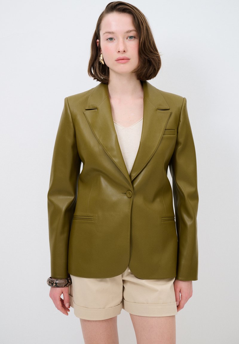 Blazer in pelle sintetica verde oliva con collo a scollo e chiusura a un bottone. Presenta due tasche frontali e una vestibilità sartoriale. Indossato sopra una maglietta beige.