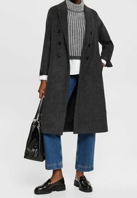Cappotto lungo grigio in misto lana con design doppiopetto, caratterizzato da bottoni neri e spalle strutturate, indossato sopra un maglione a coste e jeans.