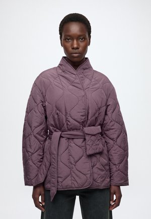 Femme portant une veste matelassée violette avec une ceinture et une petite pochette attachée, debout devant un fond clair uni.