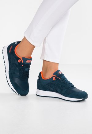 Nohy v bílých kalhotách a námořnicky modrých teniskách značky ASICS s oranžovými detaily, stojící na bílém podlaze před jednobarevným pozadím.