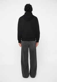 Vyras su garbanotais plaukais, dėvintis juodą džemperį su kapišonu ir plačius tamsiai pilkus džinsus, stovi atsukęs nugara prieš baltą foną.