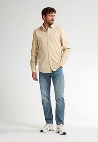 Beige langærmet skjorte med knappet krave, tekstureret stof, kombineret med lyseblå jeans og hvide sportssko.