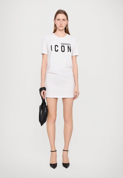 T-shirt en coton blanc avec imprimé noir "ICON", associé à une jupe blanche ajustée. Modèle tenant un sac en cuir noir, portant des talons noirs avec des brides à la cheville.