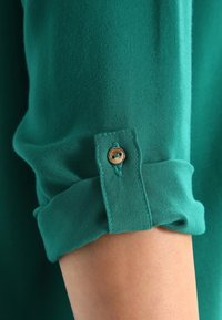 Camicia in cotone verde con maniche arrotolate e dettaglio in legno, caratterizzata da una texture liscia e da una leggera lucentezza.