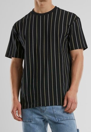 Homme portant une chemise noire à manches courtes avec de fines rayures verticales blanches et jaunes et un jean en denim bleu clair.