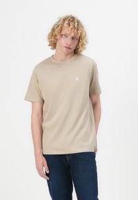 T-shirt en coton beige avec un col rond, des manches courtes et un petit logo triangulaire sur la poitrine gauche. Porté avec un jean bleu foncé.