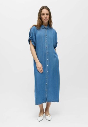 Femme portant une robe midi en jean bleue boutonnée sur le devant, avec des manches courtes retroussées et des ballerines blanches pointues, debout sur un fond blanc uni.