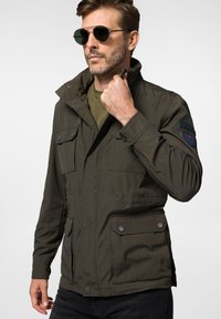 Oliwkowa kurtka zapinana na zamek z wysokim kołnierzem, wieloma przednimi kieszeniami i zapięciami na zatrzask. Tkanina ma gładką, lekką fakturę.