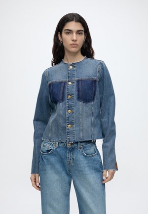Kvinde med langt mørkt hår bærer en knappet, kraveløs denimjakke og matchende blå jeans, stående mod en ensfarvet lys baggrund.