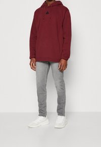 Viininpunainen huppari mustalla kolmiomerkillä, yhdistettynä vaaleanharmaisiin slim-fit-farkkuihin ja valkoisiin lenkkareihin yksinkertaisella taustalla.