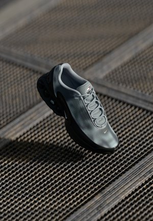Grauer Sportschuh mit Netzstoff, runder Spitze und auffälliger Luftdämpfungssohle. Verfügt über eine Schnürung vorne und markante Logos.