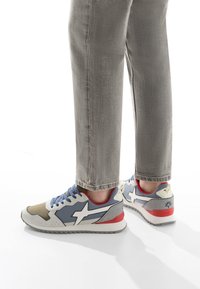 Scarpe sportive con una parte superiore grigio chiaro, blu e beige con accenti bianchi, materiali texturizzati e un tacco rosso. Design con stringhe e suola con motivo.