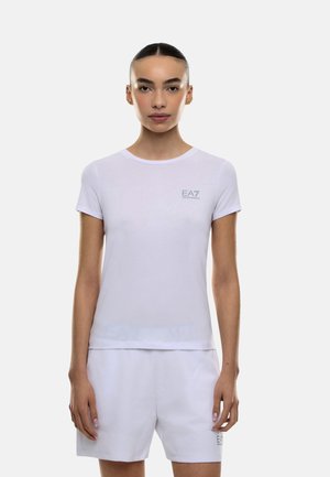 Nainen, jolla on taaksepäin kammatut hiukset, pukeutuneena valkoiseen EA7 Emporio Armani -merkkiseen lyhythihaiseen t-paitaan ja vastaaviin valkoisiin shortseihin, seisomassa yksinkertaisella taustalla.