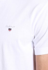 Vit kortärmad t-shirt i bomull med en liten broderad "GANT"-logotyp och sköldemblem på vänster bröst.