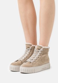 Světle hnědé semišové vysoké tenisky s bílým kožešinovým podšívkou, bílými tkaničkami a silnými bílými gumovými podrážkami, nošené na holých nohách.