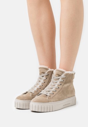 Jasnobrązowe zamszowe sneakersy typu high-top z białym futerkiem w środku, białymi sznurówkami i grubą białą gumową podeszwą, noszone na golych nogach.