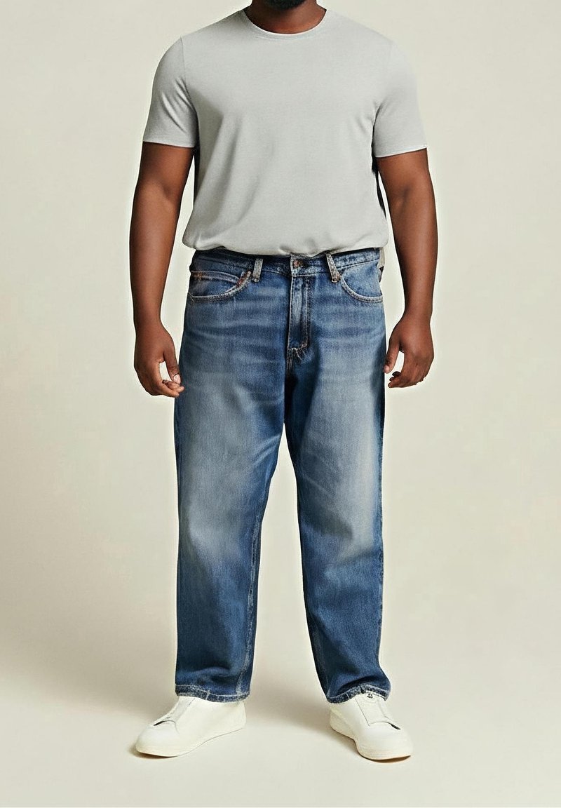 Homme portant un t-shirt gris clair, un jean bleu ample et des baskets blanches sans lacets, debout devant un fond uni.
