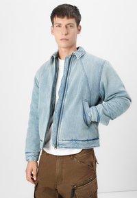 Veste en denim bleu clair avec fermeture éclair, poches latérales et détails cousus. Présente un col côtelé et un tissu texturé.