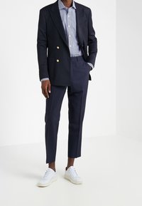 Blazer doppiopetto blu navy con bottoni dorati, abbinato a una camicia a righe e pantaloni blu navy. Sneakers bianche completano l'outfit.