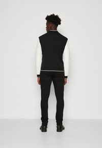Chaqueta bomber negra con mangas blancas, puños y cuello acanalados, llevada sobre pantalones negros y botas negras. Vista trasera, fondo liso.