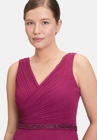 Fukszia színű ujjatlan ruha redőzött felsőrésszel és V-alakú nyakkivágással. Gyöngyös derékrésszel rendelkezik, amely texturált anyagból készült, sima befejezéssel.