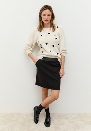 Cremefarbener Strickpullover mit schwarzen Herzen und einem rosa Herz-Detail. Schwarzer Rock mit strukturierter Optik. Schwarze Schuhe mit glänzendem Finish und gepunktete Socken.