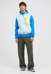 Hellblau gepolsterte Weste mit einer gelben Reißverschlusstasche, blauen Kapuzenärmeln, kombiniert mit dunkelgrünen Hosen und grauen Turnschuhen. Glattes und leichtes Material.